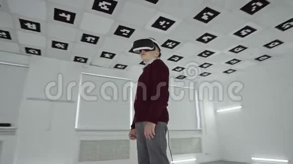 穿着棕色毛衣的小男孩在一个充满白色霓虹灯的空玩室里透过虚拟现实眼镜看着视频的预览图