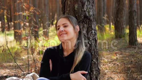 4k视频美丽的微笑女人在森林或公园的地面上放松靠在杉树上视频的预览图