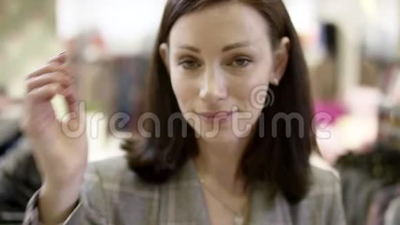 漂亮的成年女人正看着镜头微笑着抚摸着她在服装店的头发闭上脸视频的预览图