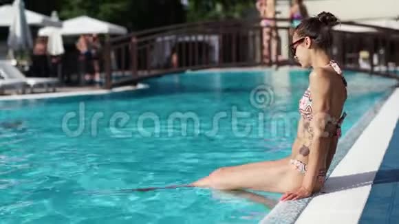 年轻的身材苗条的女人有着漂亮的平坦的腹部坐在池边把腿浸入水中享受阳光明媚的一天视频的预览图