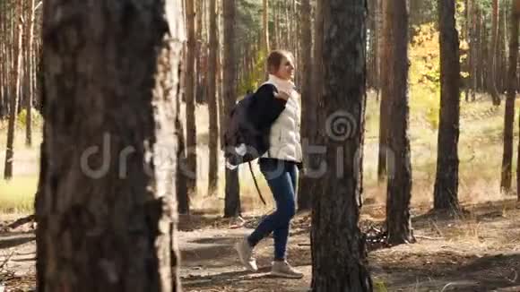 阳光明媚的秋天带着背包在森林里徒步旅行的快乐微笑年轻女子4k视频视频的预览图