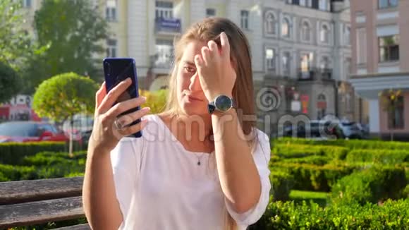 城市公园长头发的美女坐在长凳上在智能手机上自拍的4k特写视频视频的预览图
