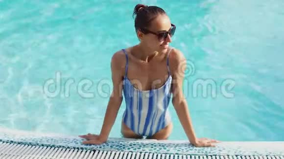 穿着泳装戴墨镜的漂亮性感运动女孩双手站在池边蓝水背景视频的预览图