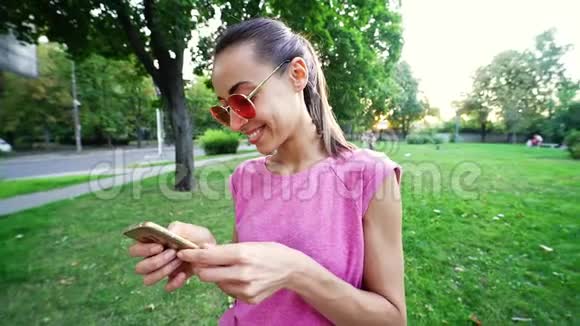 在日落城绿色公园背景下戴着粉红色眼镜的年轻微笑女子用手机慢动作视频的预览图