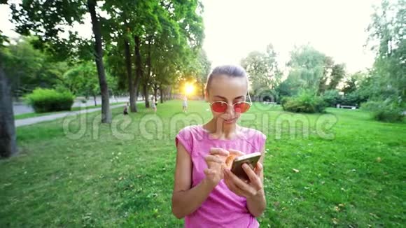 在日落城绿色公园背景下戴着粉红色眼镜的年轻微笑女子用手机慢动作视频的预览图