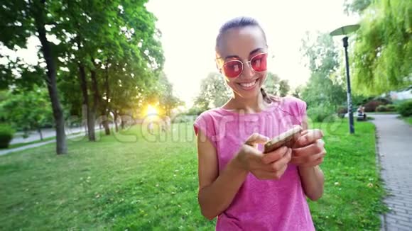 戴着粉红色眼镜的年轻微笑女子站在日落城绿色公园的背景上用手机视频的预览图