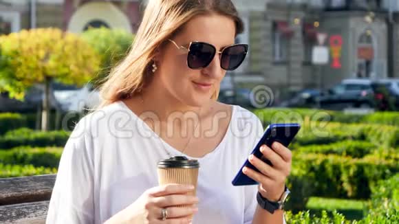 4k视频微笑的年轻女性长发在公园里喝杯咖啡在风和日丽的时候使用智能手机视频的预览图