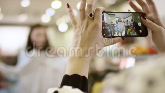 年轻快乐的女人正在试穿衬衫而另一个女人正在服装店的智能手机上拍照视频的预览图