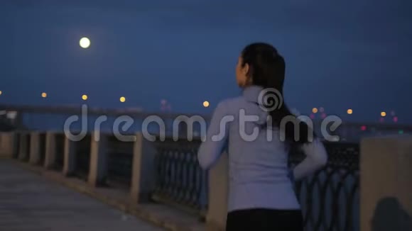 夜间慢跑一个穿着运动服的女孩在一个夜晚城市的背景下沿着一条荒芜的夜堤奔跑回来视频的预览图