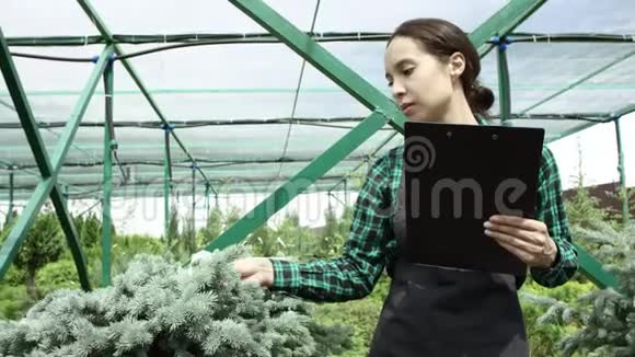 穿着工作服装和灰色围裙的布鲁内特女孩看着温室里的植物做笔记视频的预览图