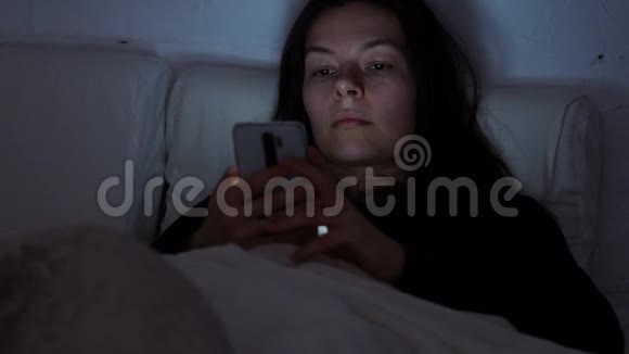晚上躺在床上的年轻女人使用智能手机社交网络和互联网滚动视频的预览图