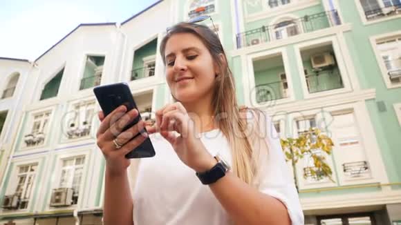 美丽的微笑女孩站在城市街道上使用智能手机的特写低角度视频视频的预览图