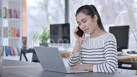 带手提电脑的年轻亚洲女人在办公室里用智能手机说话视频的预览图