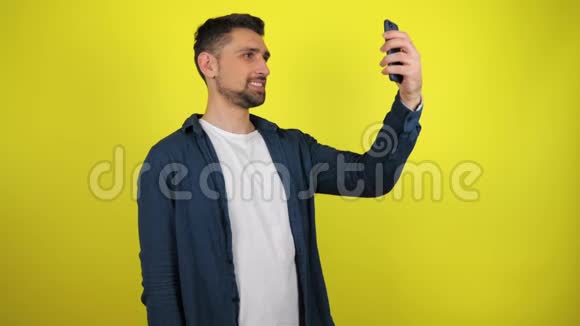一个穿着蓝色衬衫和白色T恤的年轻人在智能手机上自拍视频的预览图