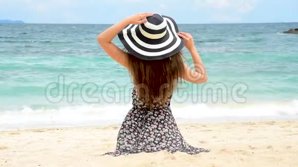 夏天棕褐色的女人在海滩上度假欢快的女人穿着夏装戴着草帽坐在沙滩上眺望大海提姆视频的预览图