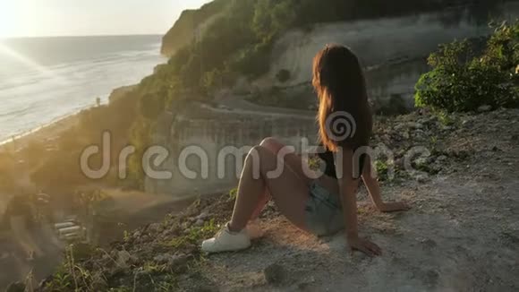 年轻的女孩坐在岩石的背景上享受着大自然的美丽女人看着海洋日落背景视频的预览图