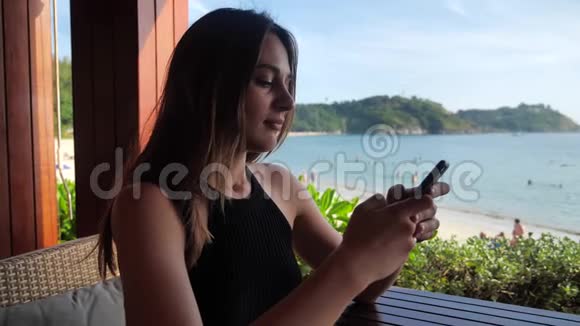 美丽的女人坐在咖啡馆里点击靠近大海的海滩的电话背景是4K视频的预览图