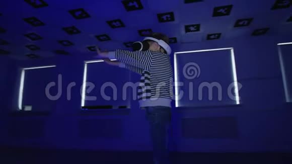 一个穿着vr耳机的小男孩在一个充满闪烁的霓虹灯的空玩室里挥舞着双手追踪弧光射击360视频的预览图