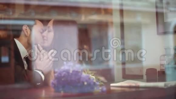 年轻的男人和漂亮的黑发欧洲女人穿着白色连衣裙在窗边的咖啡馆里热情地接吻视频的预览图