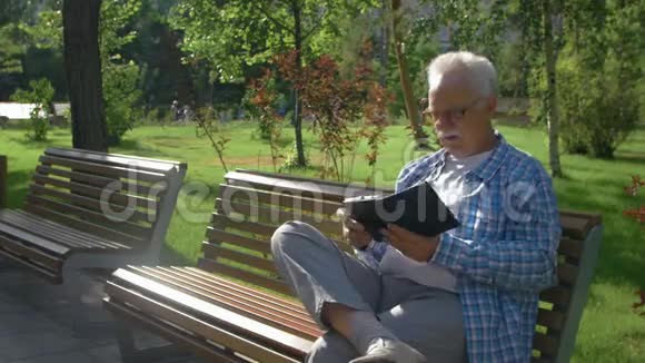 一位戴眼镜的老人在公园长椅上看电子书视频的预览图