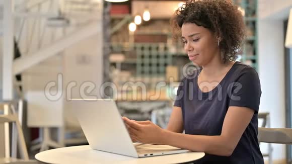 带手提电脑的年轻非洲女人在咖啡厅里用智能手机说话视频的预览图