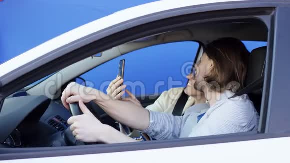 Brunette男子和金发女子正在白色汽车内拍摄蓝色背景的照片有趣的司机和他的快乐视频的预览图