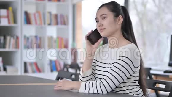 美丽的亚洲青年妇女在图书馆讲智能电话视频的预览图