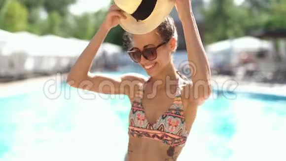 穿着比基尼帽子和太阳镜的漂亮年轻女人在游泳池的背景下快乐地笑着视频的预览图