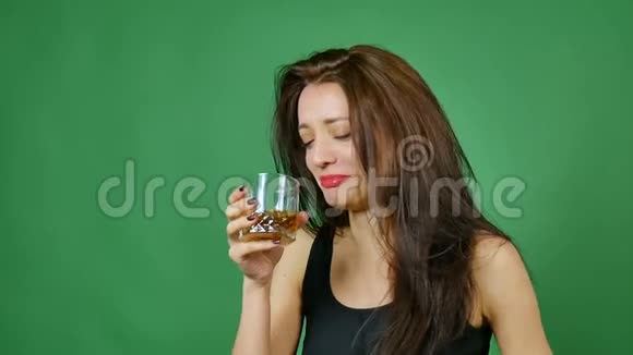 年轻的黑发悲伤的女孩正在工作室里喝着一杯绿色背景的威士忌的女人视频的预览图