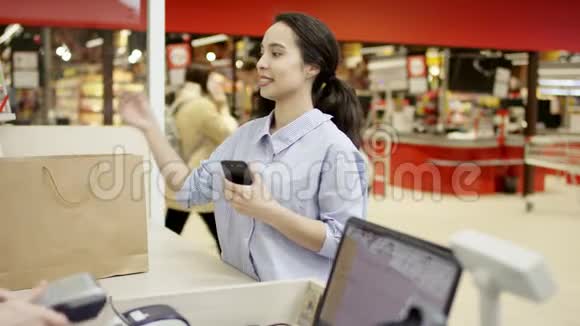 漂亮的黑发女顾客拿着智能手机谢谢收银员微笑着从收银台拿着纸购物袋视频的预览图
