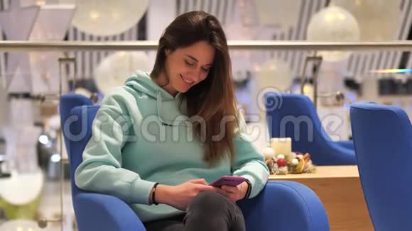 穿着绿松石运动衫的年轻女子肖像她坐在免税区点击智能手机她期待她的飞行视频的预览图