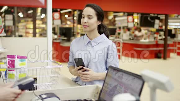 有魅力的女性顾客站在超市的收银机上通过智能手机应用程序付款视频的预览图