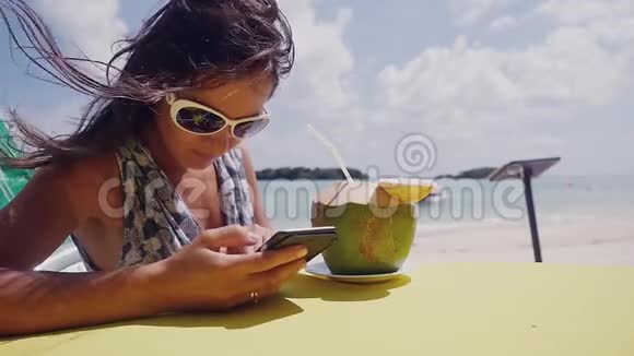 在海滩上年轻的黑发女人戴着太阳镜用手机喝着椰子做的鸡尾酒视频的预览图