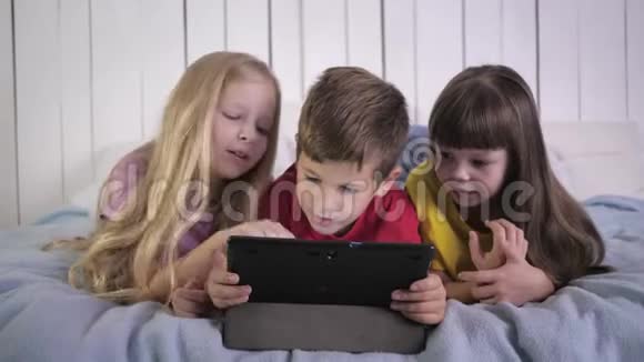 快乐的童年穿着多色t恤的小朋友在平板电脑上玩耍在家躺在床上聊天视频的预览图