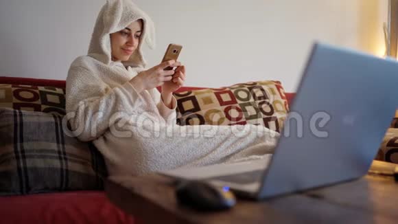 穿着有趣的小兔子睡衣坐在红色沙发上用智能手机做着快乐的年轻女人视频的预览图