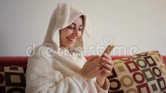 穿着滑稽小兔子睡衣坐在红色沙发上用智能手机做特写欢快的笑着的年轻女子视频的预览图