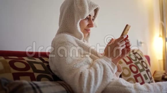 穿着滑稽的小兔子睡衣坐在红色沙发上用智能手机微笑的年轻女人视频的预览图