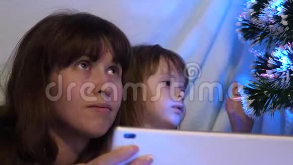 圣诞节晚上妈妈和女儿在一个带花环的帐篷里的儿童房间里在平板电脑上玩和看动画片视频的预览图