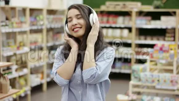 开朗的年轻白种人女人听着耳机里的音乐在超市背景下快乐地跳舞和微笑视频的预览图