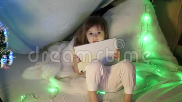 可爱的小女孩在孩子们的房间里沙发上有一台平板电脑圣诞节晚上带平板电脑的小男孩视频的预览图