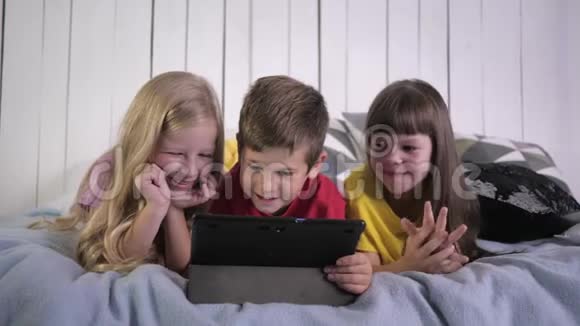现代童年时代穿着多种颜色t恤的小伙伴在家里躺在床上时使用数码平板电脑聊天视频的预览图