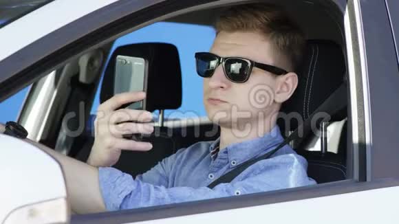 戴墨镜的年轻人正带着自拍和驾驶蓝纱背景的汽车他坐在白色的车里视频的预览图