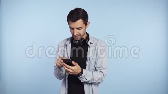 穿着蓝色t恤脖子上戴着耳机的英俊好奇的男人展示了智能手机和绿色屏幕智能手机视频的预览图