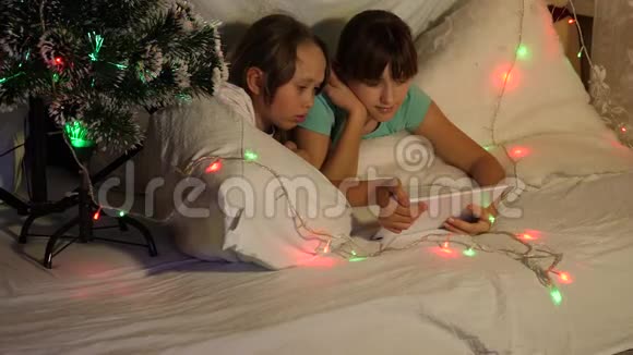 孩子们在圣诞节晚上玩平板电脑在孩子们的房间沙发花环在房间里玩平板电脑的姐妹视频的预览图