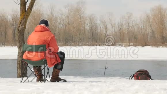 一个穿橙色夹克的人在冬天钓鱼在冰冷的河流的岸边雪中的海岸视频的预览图