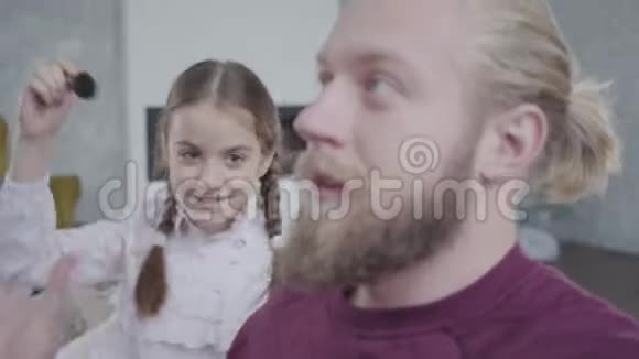可爱的白种人女孩用化妆刷梳理父亲的胡须的肖像女儿打扰疲惫的父亲重点视频的预览图