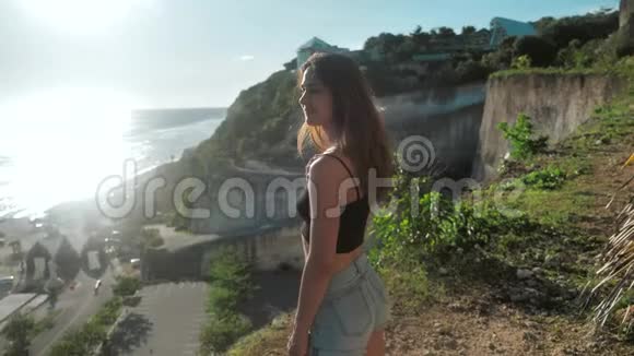 迷人的年轻女孩站在悬崖上日落她站在悬崖边上看着大海和头发视频的预览图