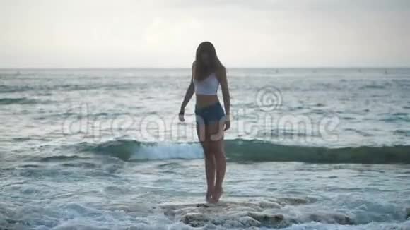 穿着牛仔短裤的女孩在石滩上散步海浪拍打着海岸4k视频的预览图
