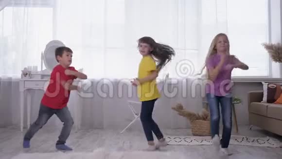 孩子们在家里度假时穿着多种颜色的T恤衫在白色房间的明亮窗户上跳舞和跳跃视频的预览图