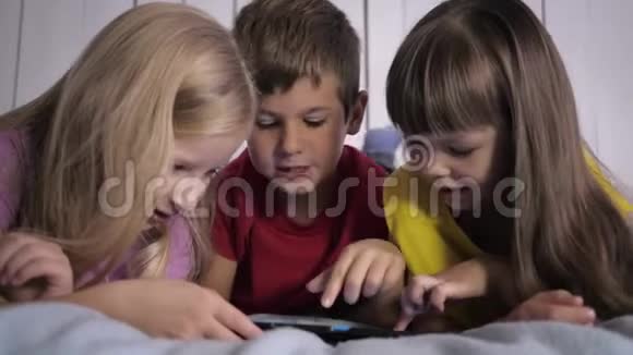 孩子们学习现代科技身穿多色t恤的小伙伴在家中躺在床上使用平板电脑视频的预览图